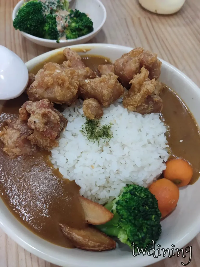 九本家唐揚雞咖哩飯包含酥炸金黃雞塊與深褐色咖哩醬汁，配料有青花菜、馬鈴薯塊與圓形紅蘿蔔，白飯灑上海苔粉，展現日式定食擺盤與濃郁醬汁質感。