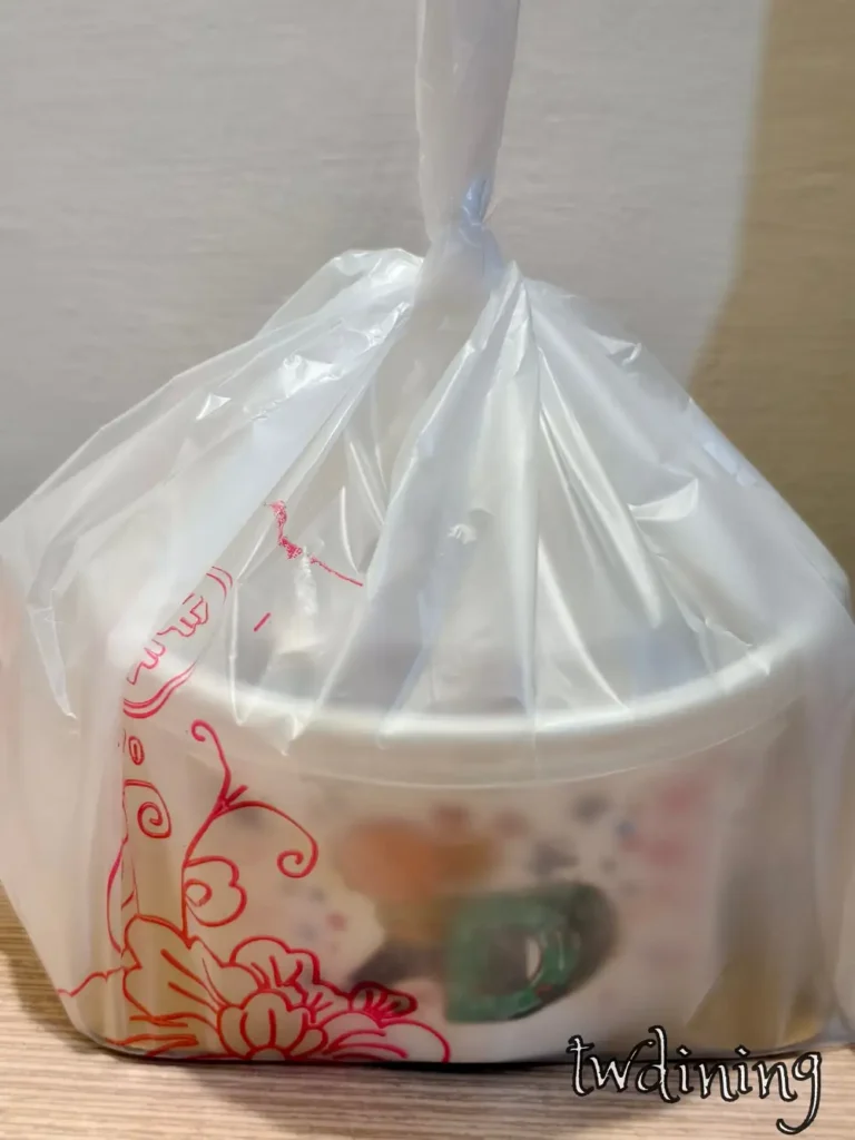 九本家日式料理外帶包裝使用白色圓形紙質餐盒，外層套用印有紅色花卉圖騰的半透明塑膠袋並打結固定，呈現台中北區餐廳外帶外送服務的打包視覺。