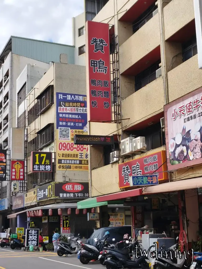 北平路贊鴨餐廳外部招牌。紅色長型直式看板標示鴨肉飯麵與快炒，結合台中北平路熱鬧街景、機車群、店面廣告與周邊店家遮雨棚，提供地理位置定位與用餐環境街拍視覺。